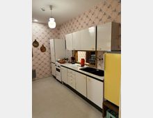 Küche im Retro-Stil mit weißen Schränken, einem gelben Kühlschrank und einer Arbeitsplatte mit Spüle und Herd. Die Wände sind mit gemusterten Tapeten verziert, und eine runde Hängelampe beleuchtet den Raum.