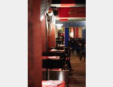 Ein gemütlicher Innenraum eines Restaurants mit dunklen Holzmöbeln, roten Tischoberflächen und dekorativen Stoffbahnen an der Decke. Die Wände sind mit gemusterten Tapeten gestaltet, und im Hintergrund sind diffus beleuchtete Bereiche zu sehen.