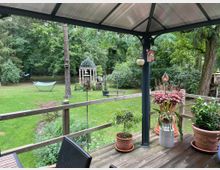 Eine überdachte Holzterrasse mit Topfpflanzen und dekorativen Elementen, die in einen großzügigen, begrünten Garten mit einer Hängematte, einem Pavillon aus Metall und hohen Bäumen führt. Im Hintergrund ist dichter Wald zu sehen.