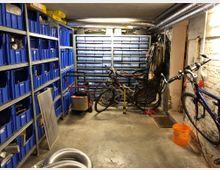 Ein Kellerraum mit Regalen voller blauer Plastikboxen auf der linken Seite und Schubladenschränken an der Stirnseite. Mehrere Fahrräder stehen oder hängen an der rechten Seite, und es befindet sich weitere Lagerausstattung wie Kabelrollen und Werkzeuge im Raum.