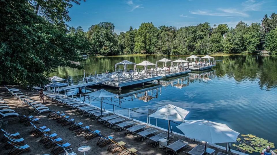 A lakeside recreational area featuring a sandy beach with sun loungers, tables, and white umbrellas. Floating docks with shaded seating extend over the calm water, surrounded by dense green trees under a clear blue sky.