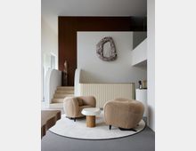 Ein moderner Wohnzimmerbereich mit zwei beigefarbenen, gepolsterten Sesseln auf einem runden, weißen Teppich. Ein kleiner Tisch mit Holzfuß und weißer Tischplatte steht zwischen den Sesseln, im Hintergrund befinden sich eine geschwungene Treppe, eine Strukturwand mit Holzverkleidung und ein abstrakter Wandspiegel.