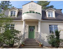 Zweistöckiges Wohnhaus mit hellem Putz und einem Walmdach mit Dachgauben. Die Fassade zeigt eine grüne Haustür mit halbrundem Glasfenster, einen kleinen Balkon mit Geländer und Säulen sowie umliegende Pflanzen und einen Treppenaufgang aus Ziegelsteinen.