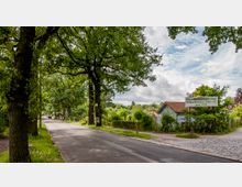 Straße mit großen, dicht belaubten Bäumen auf beiden Seiten; rechts ein Schild mit der Aufschrift „Dauerkleingartenanlage Schlachtensee-Süd e.V. gegr. 1919“ und ein kleines Gartengebäude mit rotem Dach. Im Hintergrund sind Gärten und grüne Landschaft erkennbar.