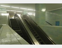 Untergrundstation mit zwei Rolltreppen, flankiert von Wänden und Säulen, die mit hellgrünen und blauen Kacheln verkleidet sind. Die Decke ist mit langen Leuchtstoffröhren beleuchtet.