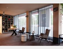 Ein moderner, gemütlicher Innenraum mit großen, bodentiefen Fenstern, durch die lichtdurchlässige Vorhänge hängen. Die Einrichtung umfasst einen runden Holztisch mit Topfpflanzen, gepolsterte Sessel, kleine Hocker und einen Beistelltisch. Im Hintergrund steht ein offenes Regal mit dekorativen Gegenständen und dem Schriftzug "VOLKSPARK".