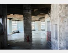 Unfertiger Innenraum eines Gebäudes mit freigelegter Betonstruktur, teilweise verputzten Wänden und einem Boden aus hellen Fliesen. Große Fenster durchfluten den Raum mit Tageslicht und bieten Ausblick auf eine Stadtlandschaft.