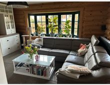 Gemütliches Wohnzimmer mit Holzwänden und großem Ecksofa aus grauem Leder. Im Vordergrund steht ein weißer Couchtisch mit Büchern und einer Vase mit Tulpen, während im Hintergrund ein Fenster mit grünen Rahmen und zahlreichen Zimmerpflanzen zu sehen ist.