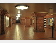 U-Bahn-Unterführung mit roten Backsteinwänden, quadratischen Säulen und einem Boden aus roten Fliesen. Die Decke ist mit runden Deckenleuchten ausgestattet, und an den Wänden befinden sich Karten und Hinweistafeln.