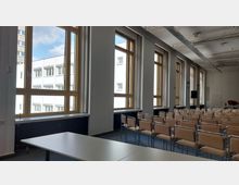 Ein moderner Seminarraum mit hohen Holzrahmenfenstern, die Tageslicht hereinlassen, Reihen von beigefarbenen Stühlen mit Metallrahmen und einem weißen Tisch im Vordergrund. Die Decke ist mit sichtbaren technischen Installationen ausgestattet, und im Hintergrund befindet sich ein Lautsprecher sowie eine rote Sitzgelegenheit.