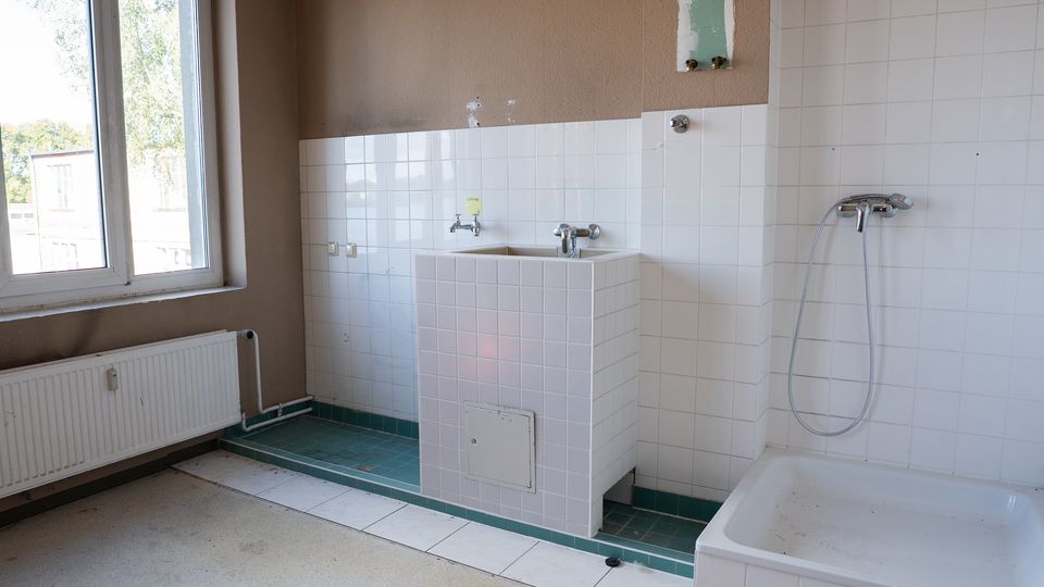 Unrenovierter Sanitärraum mit weiß gefliesten Wänden, einem gefliesten Waschbeckenblock und einer Duschwanne mit Handbrause. Links befindet sich ein großes Fenster über einem Heizkörper; der Boden ist teils gefliest und teils roh, mit sichtbaren Installationsspuren an der Wand.