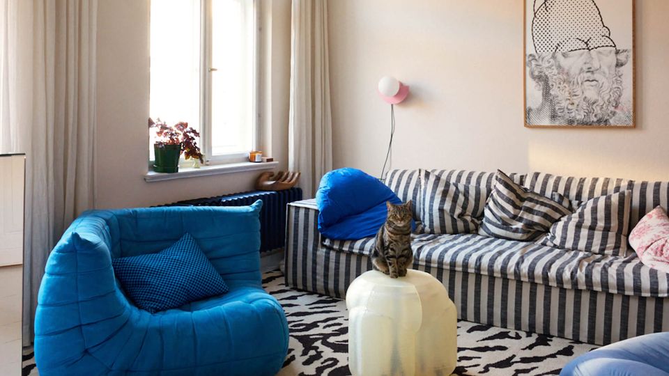 Bright living room with tall windows and light curtains, featuring a striped sofa, a blue lounge chair, and a translucent round coffee table on a black-and-white patterned rug. A wall sconce and framed artwork hang above the sofa, with a radiator and potted plant beneath the window.