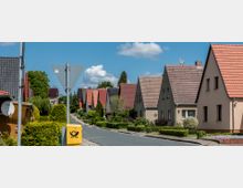 Straße mit Reihen von Einfamilienhäusern, gekennzeichnet durch rote, braune und schwarze Satteldächer sowie gepflegte Vorgärten mit Hecken und Sträuchern; im Vordergrund ein gelber Postkasten und ein Verkehrsschild.