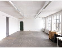 Weißer, leerer Raum mit einer einzigen grünen Tür an der Rückwand, großen Fenstern auf der rechten Seite, die Tageslicht hereinlassen, und einem Betonboden. An der Seite stehen einige Holzkisten und Möbel, während die Decke mit schlichten Leuchtstoffröhren ausgestattet ist.