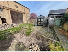 Ein Garten mit einer Mischung aus Rasen- und gepflasterten Flächen, im Vordergrund sind Beete mit jungen Sträuchern. Im Hintergrund befinden sich eine große Backsteinfassade mit Holzscheunentor, ein Gewächshaus aus Glas und ein überdachter Schuppen. Ein Hund steht rechts auf dem gepflasterten Weg.