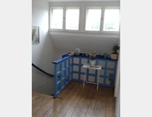 Treppenhaus mit Holzfußboden und einem hellen Geländer in blau-weißer Gitteroptik. Oberhalb des Geländers befinden sich mehrere Fenster, und auf einem kleinen weißen Tisch stehen Dekorationsgegenstände und eine Topfpflanze.