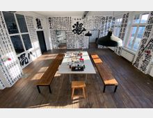 Ein heller Raum mit Holzdielenboden, weißen Wänden, die vollständig mit schwarzer asiatischer Kalligrafie bedeckt sind. Im Zentrum steht ein weißer Tisch mit Geschirr und Dekoration, flankiert von Holzbänken und beleuchtet durch große Fenster und schwarze Hängelampen.