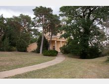 Ein Rundbau aus beigem Stein mit Säuleneingang, teilweise von Bäumen und Sträuchern umgeben, steht in einer parkähnlichen Landschaft mit Rasenfläche und geschwungenem Kiesweg.