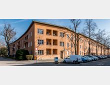 Wohnblock in einem orangefarbenen Gebäude mit drei Etagen, symmetrischen Fenstern und kleinen Balkons. Vor dem Gebäude befinden sich Bäume sowie geparkte Autos entlang der Straße.