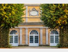 Gelb gestrichene historische Fassade mit drei großen weißen Türbögen, verziert mit halbkreisförmigen Fenstern, flankiert von zwei symmetrisch angeordneten grünen Bäumen. Oberhalb der Türen befindet sich ein ovales Fenster mit steinernen Ornamenten.