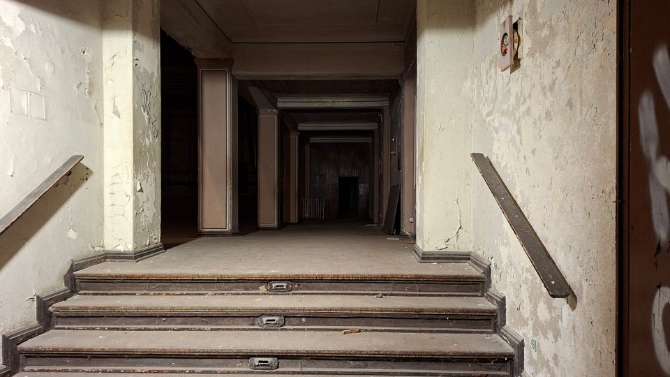 Blick von einer kurzen Treppe in einen verlassenen, langen Flur mit abblätternden Wänden, Handläufen und mehreren Stützenpfeilern. Der Raum wirkt staubig und dunkel, mit niedriger Decke und einem fernen Durchgang am Ende des Korridors.