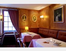 Gemütlicher Speiseraum mit holzvertäfelten Wänden, an denen Porträtgemälde und ein Stillleben hängen. Der Raum ist mit gedeckten Tischen, gepolsterten Bänken, einem beleuchteten Fenster mit Vorhängen und einer Tischlampe versehen.