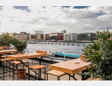 Eine Terrasse mit Holztischen und Hockern, geschmückt mit Pflanzen in Töpfen, bietet Blick auf einen Fluss und moderne Gebäude im Hintergrund. Eine Brücke und ein angrenzender Pool sind ebenfalls sichtbar.