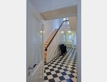 Ein Innenraum mit schwarz-weiß-kariertem Boden, einer Holztreppe mit weißem Geländer und einem Treppenaufgang. Im Hintergrund befinden sich große Fenster mit Sprossen und Heizkörper, daneben stehen zwei schwarze Stühle.