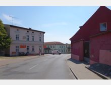 Straßenszene in einer kleinen Stadt mit mehreren Gebäuden: Links ein zweistöckiges, cremefarbenes Haus mit Schildern und Eingängen zu lokalen Geschäften; rechts ein rosafarbenes Gebäude mit einem großen Tor. In der Mitte eine asphaltierte Straße mit einem geparkten Lieferwagen und einem Fußgänger.