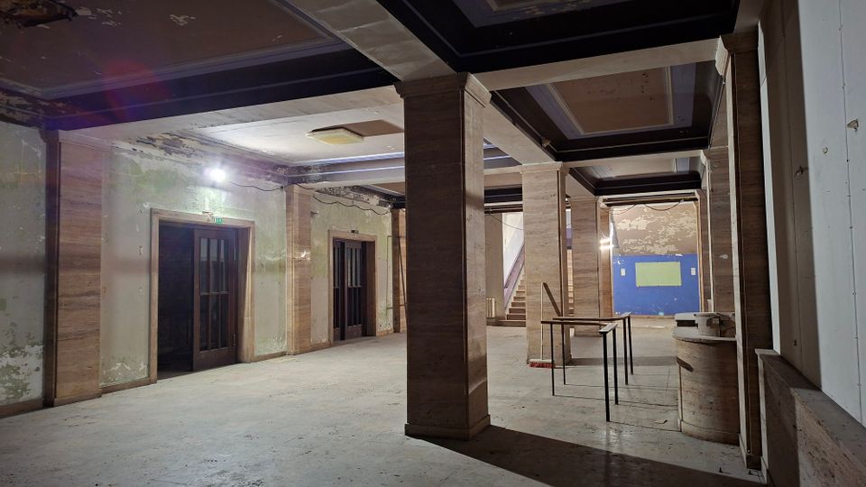 Große, unrenovierte Innenhalle eines älteren Gebäudes mit breiten Säulen, dunklen Deckenbalken und abblätterndem Putz an den Wänden. Links befinden sich doppelflügelige Türen, im Hintergrund eine Treppe, und rechts stehen provisorische Tische sowie Baumaterialien.