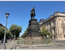 Reiterdenkmal auf einem hohen Sockel mit detaillierten Figuren, umgeben von einem grünen Zaun. Im Hintergrund eine historische Sandsteinfassade mit Fenstern und Verzierungen sowie eine von Bäumen gesäumte städtische Straße.