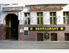 Straßenansicht eines Restaurants und einer Hotel-Pension mit den Schildern „Grossbeeren-Keller“, „König-Pilsener“ und „Restaurant“. Die Fassade ist teilweise mit dunklen braunen Fliesen verkleidet, und es gibt mehrere Fenster sowie eine Eingangspassage mit einem Tor. Ein Kreidetafel-Menü und ein beleuchtetes Werbeschild sind ebenfalls sichtbar.