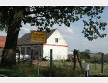 Ortseingangsschild von „Treppeln, Gemeinde Neuzelle, Landkreis Oder-Spree“ vor einem großen Baum; im Hintergrund ein Gebäude mit der Aufschrift „KFZ-Werkstatt“ sowie eingezäunte Fahrzeuge und landwirtschaftliches Gerät.