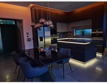 Moderne Küche mit dunklen Schränken, einer zentralen Kücheninsel mit LED-Beleuchtung und einem großen schwarzen Esstisch mit gepolsterten Stühlen. Hängende Kugelleuchten aus Metall und ein großes Fenster mit Blick ins Grüne ergänzen das elegante Design.