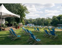 Grüne Wiese mit blauen Liegestühlen und Holztischen, die vor einer kleinen überdachten Bühne stehen, umgeben von Bäumen. Im Hintergrund fließt ein Fluss mit einem weißen Boot, dahinter eine Waldlandschaft.