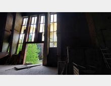 Innenraum eines verlassenen Industriegebäudes mit hohen Fensterfronten aus Glas und Stahl, durch die Tageslicht eindringt. Im Vordergrund sind Backsteinwände, eine Leiter und Sperrmüll zu sehen, während im Hintergrund ein grüner Baum sichtbar ist.