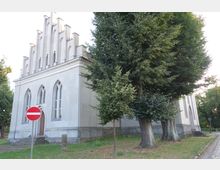 Kirchengebäude im neugotischen Stil mit spitzbogigen Fenstern und einer markanten, gestuften Fassade. Im Vordergrund steht ein Verbotsschild sowie mehrere Bäume auf einer Rasenfläche.