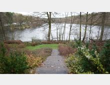 Ein Garten mit einem steinernen Weg, der zu einem Holzzaun und einem See führt. Im Hintergrund sind kahler Baumbestand, ein Bootssteg und eine ruhige Uferlandschaft erkennbar.