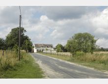 Ländliche Straße mit niedrigem, weißen Wohnhaus im Hintergrund, umgeben von Bäumen, Wiesen und Feldern; ein Strommast und Holzpfähle säumen den Weg. Der Himmel ist teilweise bewölkt.