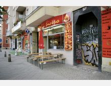 Straßenszene mit einem kleinen Bistro, das eine rote Beschilderung mit Speisenabbildungen hat. Vor dem Bistro stehen Holzbänke und Tische, ein roter Sonnenschirm ist aufgestellt; daneben ein schwarzer Hauseingang mit Graffiti und ein angrenzender Bäckerladen mit orangener Beschilderung.