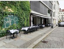 Außenbereich eines Restaurants in einer gepflasterten Straße mit mehreren gedeckten Tischen und schwarzen Stühlen vor einer Hauswand, die teilweise mit Efeu bewachsen ist. Im Hintergrund sind moderne Gebäude und parkende Autos zu sehen.