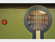 Grüne Wand mit einem großen, runden Fenster, das von einem grauen Rahmen umgeben ist und eine verzerrte, reflektierende Oberfläche zeigt. Links daneben befindet sich ein kleiner roter Kreis.