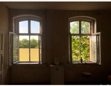 Zwei große, offene Fenster mit weißem Holzrahmen in einem schlichten, dunklen Raum. Durch die Fenster ist eine Landschaft mit Bäumen, Wiesen und klarem Himmel zu sehen.