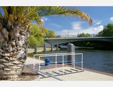 Eine Uferpromenade am Wasser mit einem Geländer und einer kleinen Rampe im Vordergrund. Im Hintergrund eine moderne Betonbrücke, grüne Bäume und ein blauer Himmel mit wenigen Wolken.