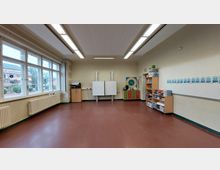 Klassenzimmer mit braunem Boden, großen Fenstern auf der linken Seite und weißen Wänden. Vorne befinden sich eine interaktive Tafel, Regale mit Materialien und ein Kalender an der Wand. Außerhalb der Fenster sind Schulgebäude und Grünflächen sichtbar.