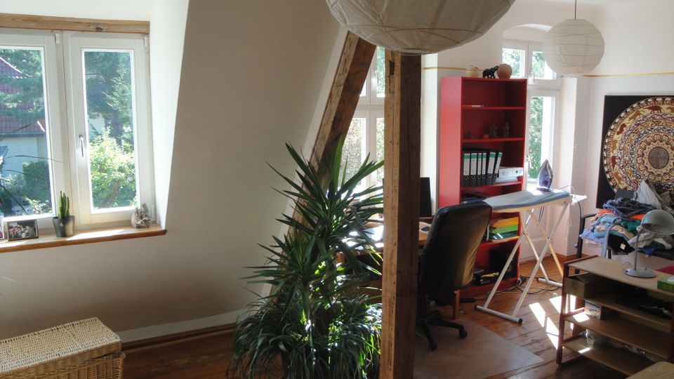 Heller Raum im Dachgeschoss mit Holzboden, großen Sprossenfenstern und sichtbaren Holzbalken, die den Raum gliedern. Zu sehen sind eine Zimmerpflanze, ein Arbeitsbereich mit Stuhl und rotem Regal sowie eine Bügelstation und niedrige Möbel.
