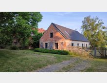 Ein ländliches Gebäude aus Ziegel- und Feldsteinen mit Satteldach und kleinen Fenstern, umgeben von einem Garten mit Bäumen, Sträuchern und einem Holzzaun. Im Vordergrund ein gepflasterter Weg und eine Rasenfläche.