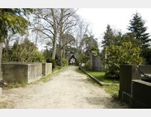 Ein friedhofsartiger Weg mit sandigem Boden, flankiert von niedrigen Mauern und dichter Vegetation. Im Hintergrund ist ein kleines hölzernes Gebäude mit einem spitz zulaufenden Dach erkennbar, umgeben von hohen Bäumen.