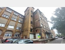 Backstein-Gebäude mit mehreren Stockwerken und stehenden, rechteckigen Fenstern; ein zentraler Turm ragt hervor. Der Eingangsbereich befindet sich auf einer erhöhten Rampe, und vor dem Gebäude sind geparkte Autos sichtbar.