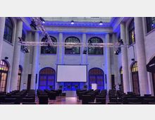 Moderner Veranstaltungsraum mit hohen Säulen, großen Bogenfenstern und einer beleuchteten Decke in Blau. Im Vordergrund stehen schwarze Stühle in Reihen, während im Hintergrund eine Projektionsleinwand, ein Podium und Stühle für eine Podiumsdiskussion zu sehen sind.